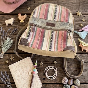 May include: A colorful striped backpack with a canvas base and leather accents. The bag features a zippered front pocket and adjustable straps. The image also includes a journal, jewelry, and decorative items, suggesting a rustic or bohemian theme.