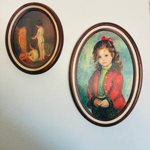 May include: Two oval framed paintings hang on a white wall. One depicts two children near a fire. The other shows a young girl in a red coat and a black hat with a red bow. The frames are dark brown with a cream-colored inner border.