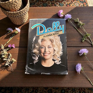 May include: A book titled "Dolly" by Alanna Nash, featuring a smiling woman with blonde hair on the cover. The title is in turquoise script. The book is on a dark wooden surface, with purple flowers and woven baskets nearby.