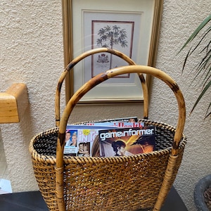 May include: Woven wicker magazine rack with bamboo handles. The rack is filled with magazines, and has a rectangular shape. The handles are curved and attached to the sides of the basket. The rack is sitting on a black surface.