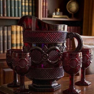 May include: A ruby red glass pitcher and matching wine glasses set on a wooden table. The pitcher has a large handle and a diamond pattern. The wine glasses have a similar design. The set is likely vintage and adds a touch of elegance.