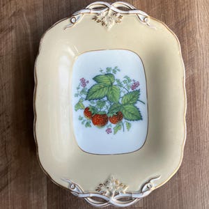 Könnte beinhalten: Ein rechteckiger, cremefarbener Keramikservierteller mit einem quadratischen Mittelteil mit Erdbeermuster. Die Erdbeeren sind rot, mit grünen Blättern und rosa Blüten. Der Teller hat einen goldenen Rand und dekorative Griffe.