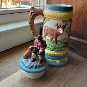 May include: A ceramic beer stein with a deer illustration and a decorative lid featuring a figurine of a hunter and dog. The stein has a wooden handle and is painted with green, yellow, and blue accents.