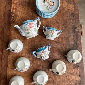 May include: A vintage miniature tea set with a floral design. The set includes a teapot, sugar bowl, cups, and saucers. The pieces are white with blue accents and colorful flower details. The set is arranged on a wooden surface.