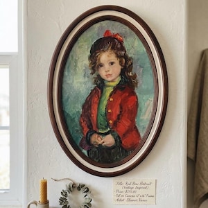 May include: An oval-framed portrait of a young child wearing a red jacket and a red bow in their hair. The painting, titled "Red Bow Portrait," is oil on canvas and is displayed on a white wall. The frame is dark brown with a cream-colored inner border.
