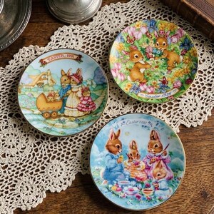 Könnte beinhalten: Drei dekorative Ostereier-Teller mit Goldrändern. Jeder Teller zeigt eine andere Szene von Hasen in Pastellfarben. Ein Teller trägt den Text "Easter 1991", ein anderer "Easter 1996" und der dritte "Easter 1999".