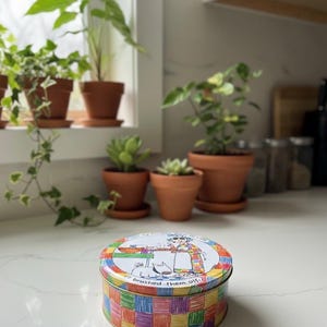 Può includere: Una scatola rotonda con un motivo a quadri colorati sui lati e un'illustrazione a cartoni animati sul coperchio. Il coperchio presenta una donna, un gatto e il testo "brushed them off". Sullo sfondo ci sono diverse piante in vaso.