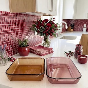 Puede incluir: Dos fuentes rectangulares de vidrio para hornear, una ámbar y otra rosa, sobre una encimera blanca. La fuente ámbar está a la izquierda y la rosa a la derecha. Una placa para salpicaduras de azulejos rojos y un ramo de bayas rojas y vegetación en el fondo.