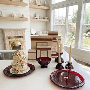 May include: A two-tiered cake with floral decorations sits on a burgundy serving platter. Other items include a burgundy bowl, candle holders, and boxes. The scene is set on a white countertop with a fireplace and shelves in the background.