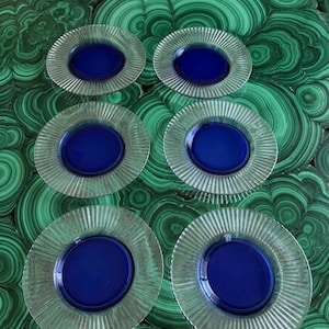 May include: Six clear glass plates with a ribbed rim and a deep blue centre. The plates are arranged on a green and black patterned surface, creating a contrast between the colours and textures.