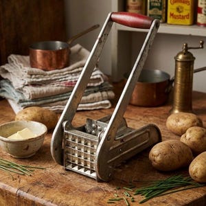 May include: A vintage potato ricer with a red wooden handle sits on a wooden surface, alongside potatoes, a bowl of butter, and chives. Copper pots and spice tins are in the background, suggesting a rustic kitchen setting.