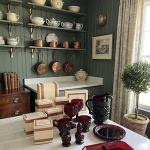May include: A kitchen interior with shelves holding vintage crockery and copper pans. A countertop displays red glassware, serving dishes, and boxes. The room has green walls, a framed picture, and a small potted tree.