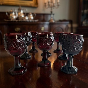 Op de afbeelding: Een verzameling robijnrode kelkglazen met sierlijke, reliëf ontwerpen. De glazen met voet zijn gerangschikt op een donkere houten tafel, die het licht reflecteert. De kelkglazen hebben een vintage uitstraling, voor een formele eetgelegenheid.