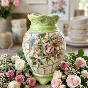 May include: A decorative vase with a mosaic design featuring floral patterns in pink and green. The vase has a light green ruffled top and base. The vase is surrounded by pink and white roses.
