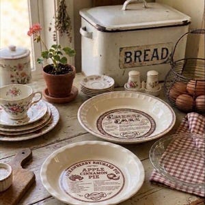May include: A white ceramic pie dish with a brown printed recipe for Strawberry Rhubarb Apple Cinnamon Pie, a matching pie dish, a white metal bread box with the word BREAD, a floral teacup and saucer, a potted plant, and a wire basket with eggs.