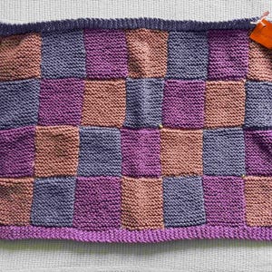 May include: A knitted blanket featuring a checkerboard design in shades of purple, mauve, and grey. The blanket has a purple border and a dark purple edge. A small orange tag is attached to the top corner.