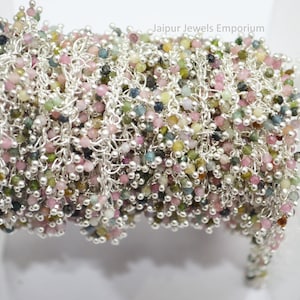May include: A close-up of a silver chain with small, multi-colored gemstone beads. The chain is coiled and the beads are a mix of pink, green, and blue.