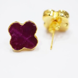 May include: A pair of gold-colored stud earrings. Each earring features a four-leaf clover design with a deep magenta center and a gold-colored border. The earrings are set against a white background.