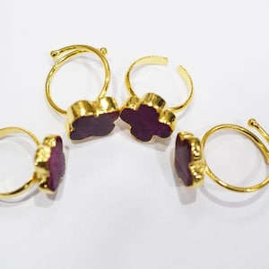 May include: Four gold-colored adjustable rings with a dark purple, clover-shaped stone setting. The rings are displayed on a white surface, showcasing the jewelry's design and color contrast.