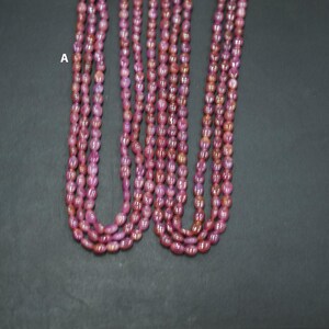 May include: Multiple strands of pinkish-red gemstone beads. The small, oval-shaped beads are strung together to form necklaces. The beads have a slightly translucent appearance, with colour variations. The background is a dark grey surface.