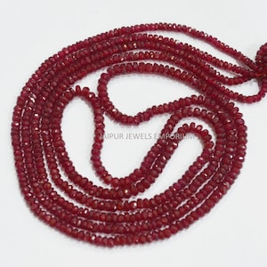 May include: A strand of red ruby beads, approximately 3 mm in size, with a faceted finish. The beads are strung on a clear thread and are arranged in a circular pattern. The beads are a deep red color and have a slight shine to them. The image shows a close-up of the beads, with the background blurred out.