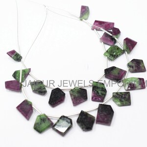 May include: A strand of green and pink gemstone beads, with a faceted, irregular shape. The beads are strung on a silver wire and are arranged in a semi-circle.