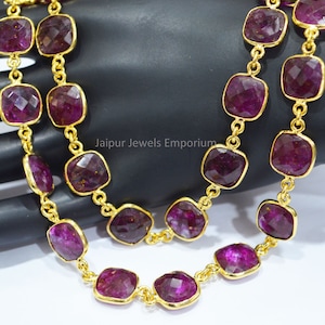 May include: A gold chain necklace with purple square-shaped gemstones. The necklace is on a hand, and the text "Jaipur Jewels Emporium" is visible.