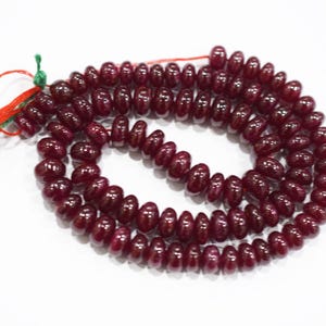 Könnte beinhalten: Eine Kette aus polierten, tiefroten Edelsteinperlen. Die Perlen sind oval geformt und zu einer durchgehenden Schlaufe aufgereiht. Ein roter und grüner Faden mit einem Anhänger ist an der Kette befestigt.