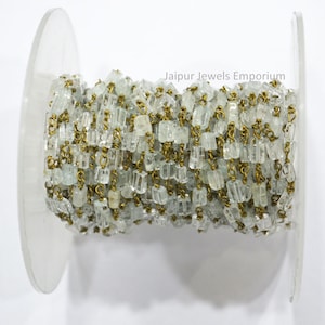May include: A spool of clear aquamarine gemstone beads strung on a brass chain. The chain is wound around a clear plastic spool. The text "Jaipur Jewels Emporium" is printed on the spool.