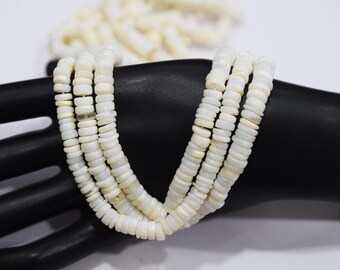 May include: A multi-strand necklace composed of small, flat, off-white beads. The necklace is draped over a black surface, with additional strands visible in the background. The beads appear to be uniform in size and shape.