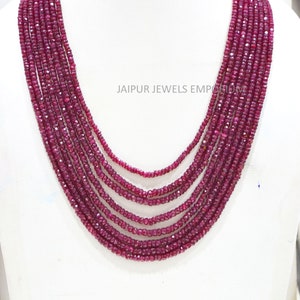 May include: A multi-strand necklace made of deep red ruby beads. The necklace is displayed on a white mannequin bust. The text "JAIPUR JEWELS EMPORIUM" is visible in the background.