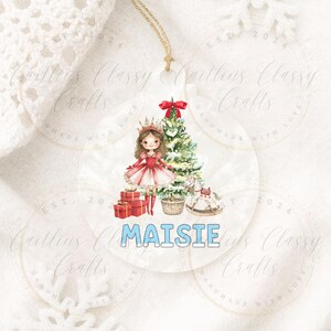 May include: A clear, round Christmas ornament featuring a princess illustration in a red dress and crown, with a Christmas tree, presents, and a rocking horse. The name "Maisie" is written in blue. A gold string is attached for hanging.