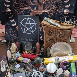 May include: A collection of spiritual and metaphysical items, including crystals, incense, a Book of Shadows, and a decorative tapestry with a moon phase design. Various crystals and gemstones are arranged on a black cloth with gold Celtic knot patterns.