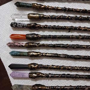 May include: A set of ten wands with different colored crystal tips. The wands are black with gold accents and have a twisted, vine-like design. The crystal tips are pink, green, clear, gray, red, pink, green, black, purple, and brown.