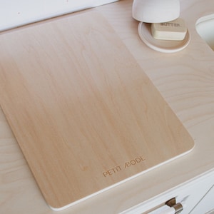 May include: A wooden toy kitchen set featuring a cutting board, butter dish, and a domed lid. The cutting board has the text "PETIT ABODE" engraved on it. The set is made of light-coloured wood and white accents.