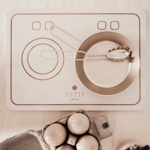 May include: A wooden play stove with two burner rings and a small pan. The stove has a logo that says "Petit Abode". There is a carton of four wooden eggs next to the stove.