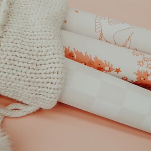May include: Three rolls of white wrapping paper with a pink and orange floral design. The rolls are stacked on top of each other and are partially covered by a white knitted fabric with a fluffy pom-pom.
