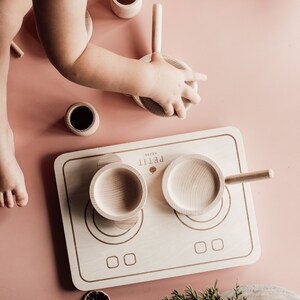 Puede incluir: Una estufa de cocina de juguete de madera con dos ollas y un mango. La estufa est&aacute; hecha de madera clara y tiene dos quemadores redondos. Las ollas tambi&eacute;n est&aacute;n hechas de madera clara y tienen mangos. La estufa est&aacute; sobre una superficie rosa.