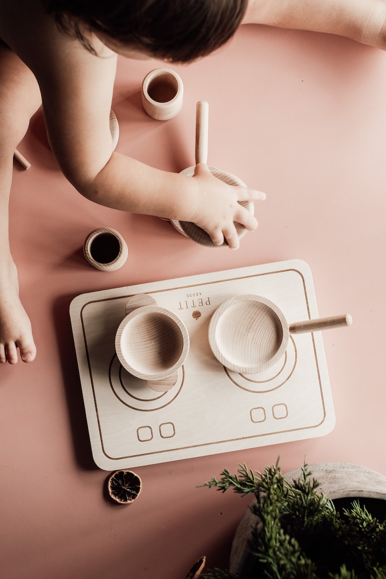 Puede incluir: Una estufa de cocina de juguete de madera con dos ollas y un mango. La estufa est&aacute; hecha de madera clara y tiene dos quemadores redondos. Las ollas tambi&eacute;n est&aacute;n hechas de madera clara y tienen mangos. La estufa est&aacute; sobre una superficie rosa.