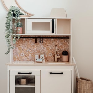 Puede incluir: Cocina de juguete blanca con un microondas negro, un grifo dorado y un salpicadero con estampado floral. La cocina tiene dos armarios con tiradores negros, uno con una puerta de cristal. Una cesta de mimbre con ruedas est&aacute; al lado de la cocina.
