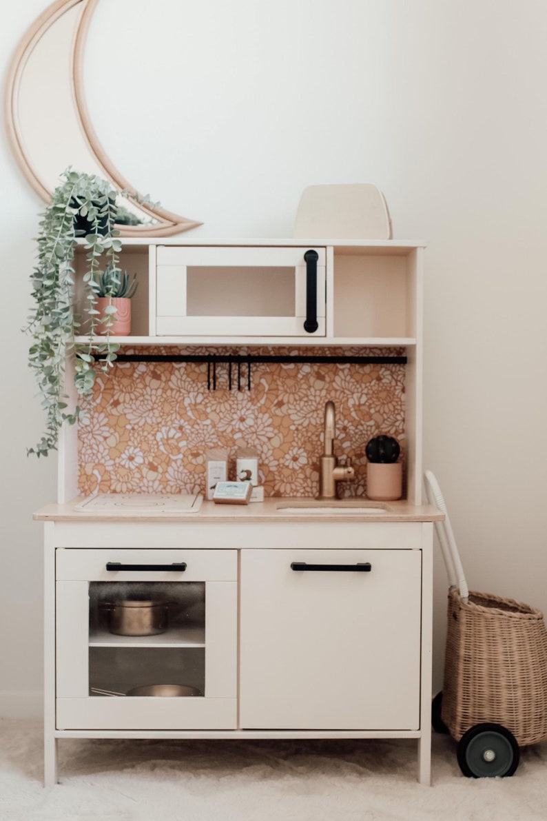 Puede incluir: Cocina de juguete blanca con un microondas negro, un grifo dorado y un salpicadero con estampado floral. La cocina tiene dos armarios con tiradores negros, uno con una puerta de cristal. Una cesta de mimbre con ruedas est&aacute; al lado de la cocina.