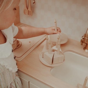 May include: A child plays with a wooden toy cake set. The set includes a cake base, three triangular cake slices, and four candles. The cake is on a wooden plate. The child is wearing a white and pink striped dress.