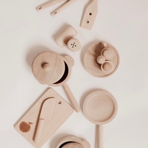 Może przedstawiać: Zestaw drewnianych zabawek do kuchni, w tym garnek z pokrywką, patelnia, szpatułka, drewniana łyżka, deska do krojenia z nożem, solniczka i talerz z trzema drewnianymi jabłkami.