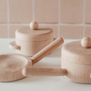 May include: A set of three wooden toy kitchen cookware, including a frying pan and two pots with lids. The cookware is made of natural wood and has a light brown colour.