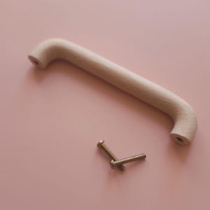 May include: A light-coloured wooden cabinet handle with a curved design, and two small screws. The handle is approximately 18 cm long. The items are set against a soft pink background, suggesting a focus on home decor or furniture.