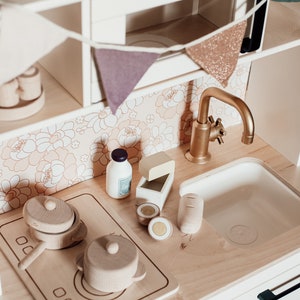 Puede incluir: Un juego de cocina de juguete de madera blanca con un grifo dorado, un fregadero de porcelana blanca y una estufa de madera con dos ollas. La cocina tiene un salpicadero con estampado floral y un estante de madera con una hilera de banderas.