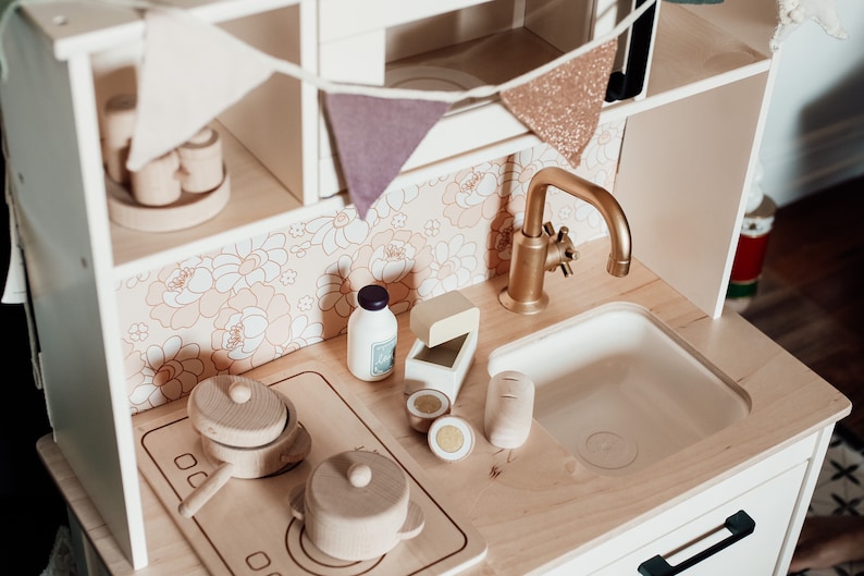 Puede incluir: Un juego de cocina de juguete de madera blanca con un grifo dorado, un fregadero de porcelana blanca y una estufa de madera con dos ollas. La cocina tiene un salpicadero con estampado floral y un estante de madera con una hilera de banderas.