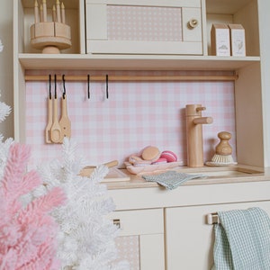May include: A white wooden toy kitchen playset with a pink and white checkered backsplash, a wooden sink, a faucet, and a wooden countertop. There are two wooden cabinets with silver handles and a green and white checkered towel hanging on one of the handles. There is a white artificial Christmas tree in the foreground.