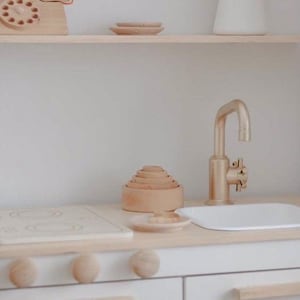 Puede incluir: Una cocina de juguete blanca con una estufa de madera, un grifo dorado y un fregadero blanco. La cocina tiene un estante de madera sobre el fregadero con un tel&eacute;fono de madera y un peque&ntilde;o plato de madera.