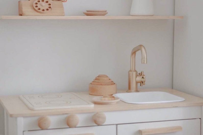 Puede incluir: Una cocina de juguete blanca con una estufa de madera, un grifo dorado y un fregadero blanco. La cocina tiene un estante de madera sobre el fregadero con un tel&eacute;fono de madera y un peque&ntilde;o plato de madera.